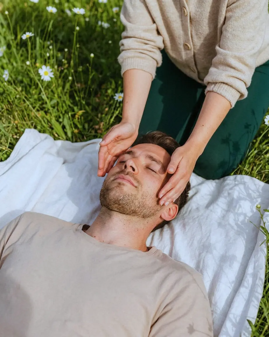 Séance de sophrologie en extérieur : les mains d'une praticienne sur les tempes d'un homme allongé dans l'herbe